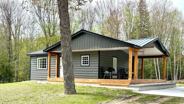 A log cabin with a covered porch and a small deck, surrounded by a grassy area and trees.