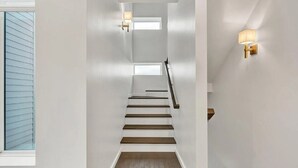 Eine Treppe mit weißen Wänden und Holzstufen, zwei Wandleuchten sowie ein Fenster mit Jalousien.