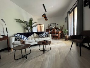 Ein modernes Wohnzimmer mit einer Couch, zwei Couchtischen, einem Esstisch mit Stühlen und einem großen Fenster mit Vorhängen.