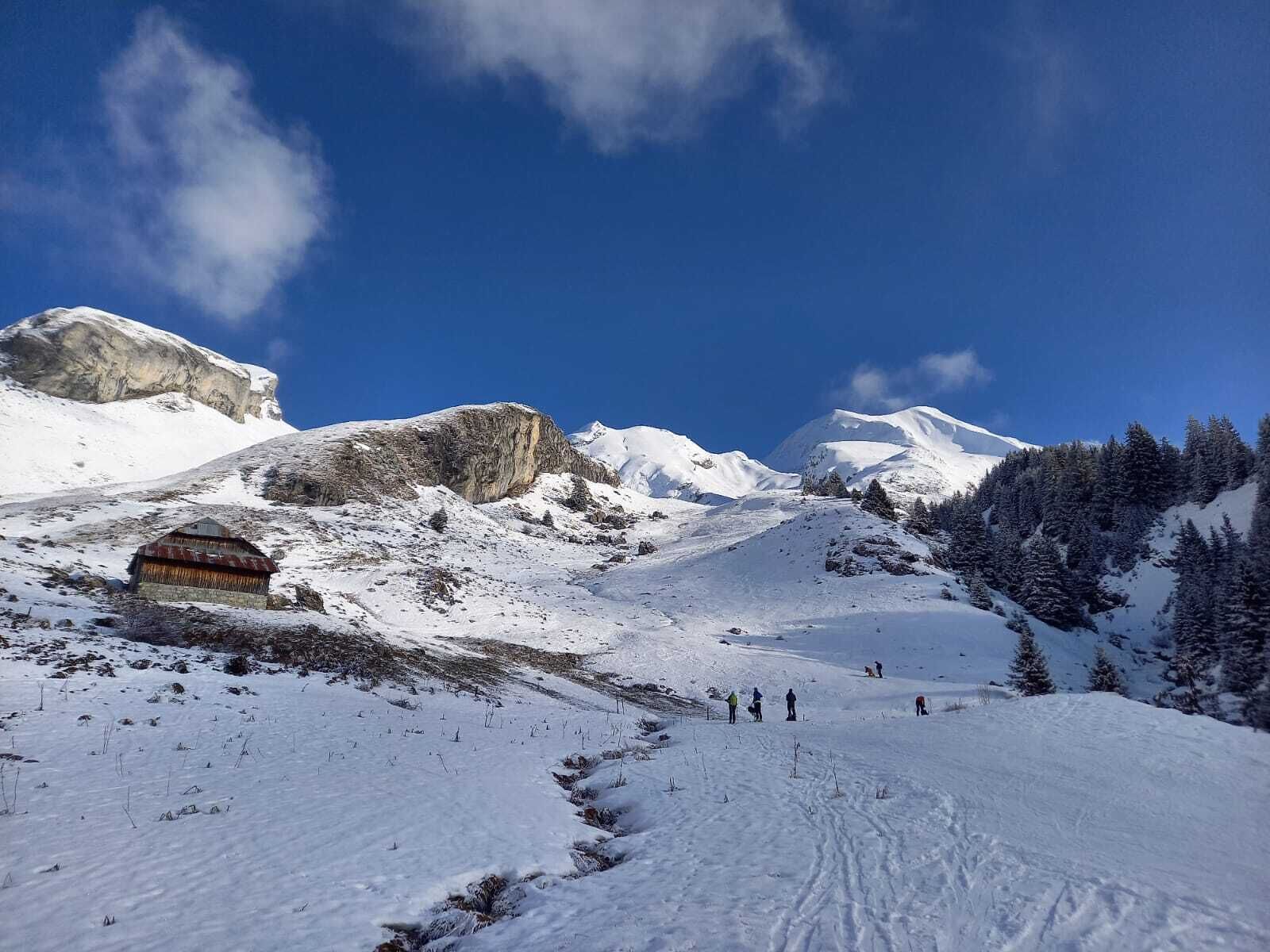 Un paysage de montagne enneigé, avec un chalet rustique, un ciel dégagé et quelques skieurs.
