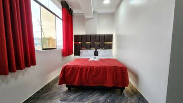 Habitación de hotel con cama, cortinas rojas, cabecero y vistas al exterior.