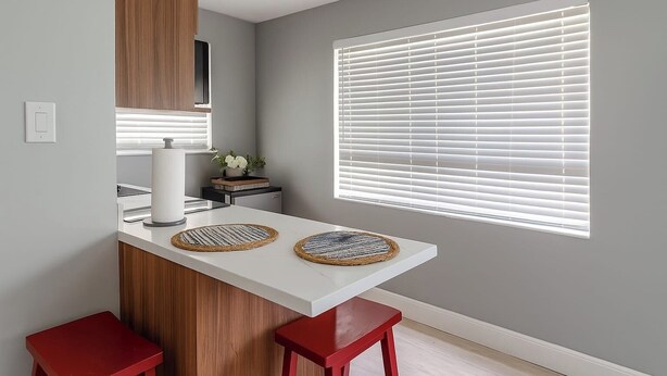 A modern kitchen with a white countertop, two red stools, a wooden cabinet, and a window with blinds.