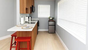 A compact kitchen with a white countertop, wooden cabinets, a small refrigerator, and a window with blinds.