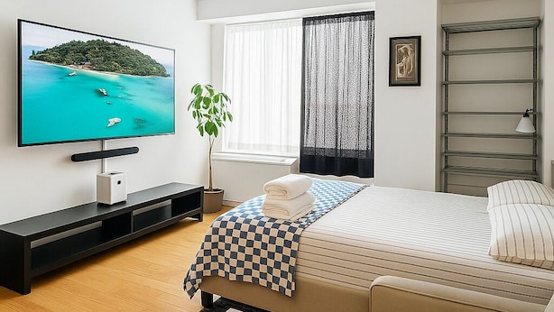 A modern hotel room with a large flat-screen TV, a bed with striped bedding, a checkered throw, a wooden coffee table, and a potted plant. - NYC Studio with Private Bathroom & Kitchenette (New York)