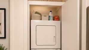 A compact laundry room with a washing machine and dryer, a small shelf with cleaning supplies, and a potted plant. - NYC Studio with Private Bathroom & Kitchenette (New York)