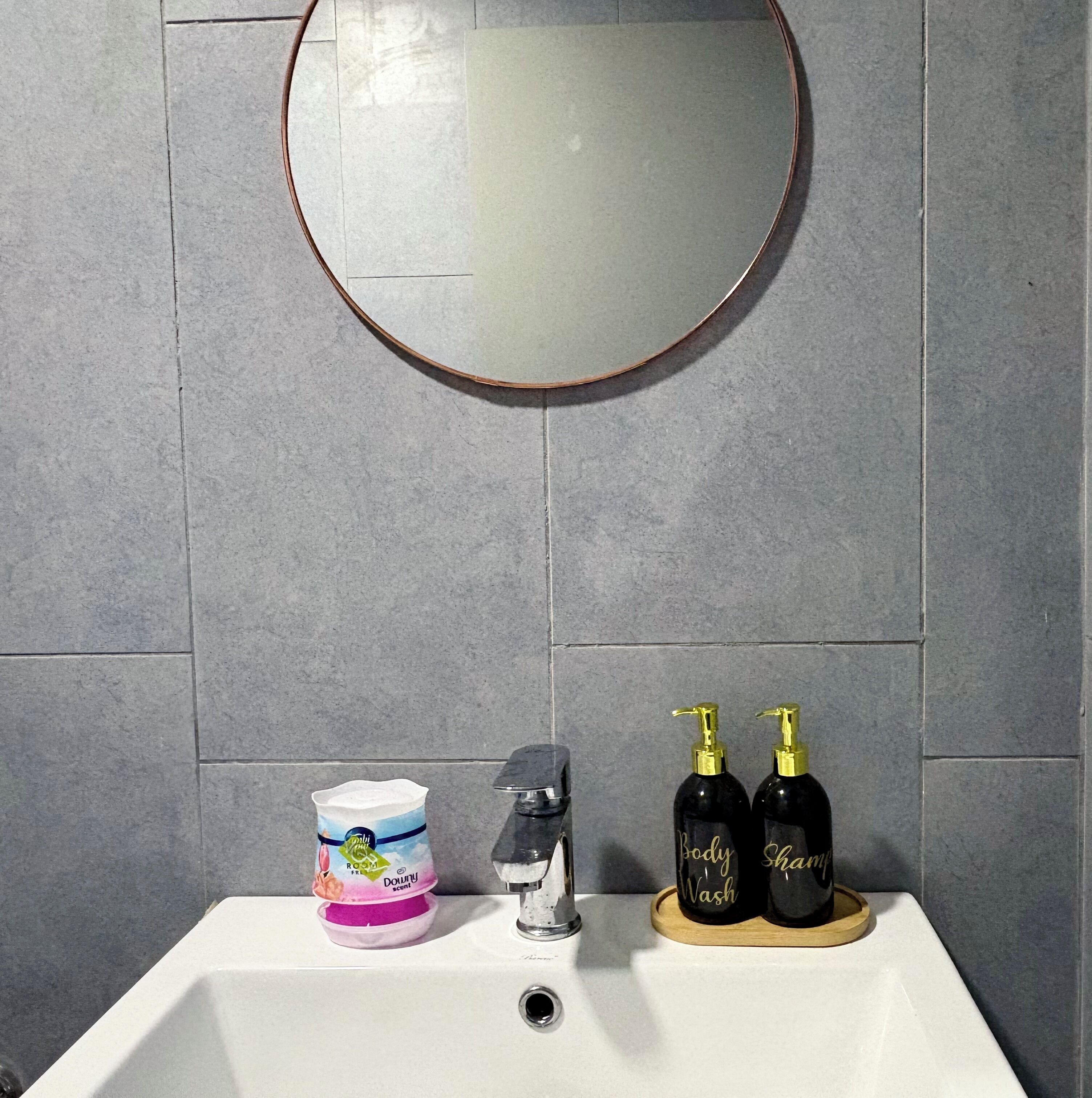 Une salle de bain avec un miroir rond, un lavabo, un distributeur de savon et un bidon d’adoucissant Downy.