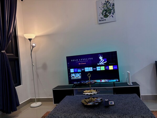 A living room with a flat-screen TV displaying a streaming service interface, a floor lamp, a decorative wall piece, and a small table with snacks.
