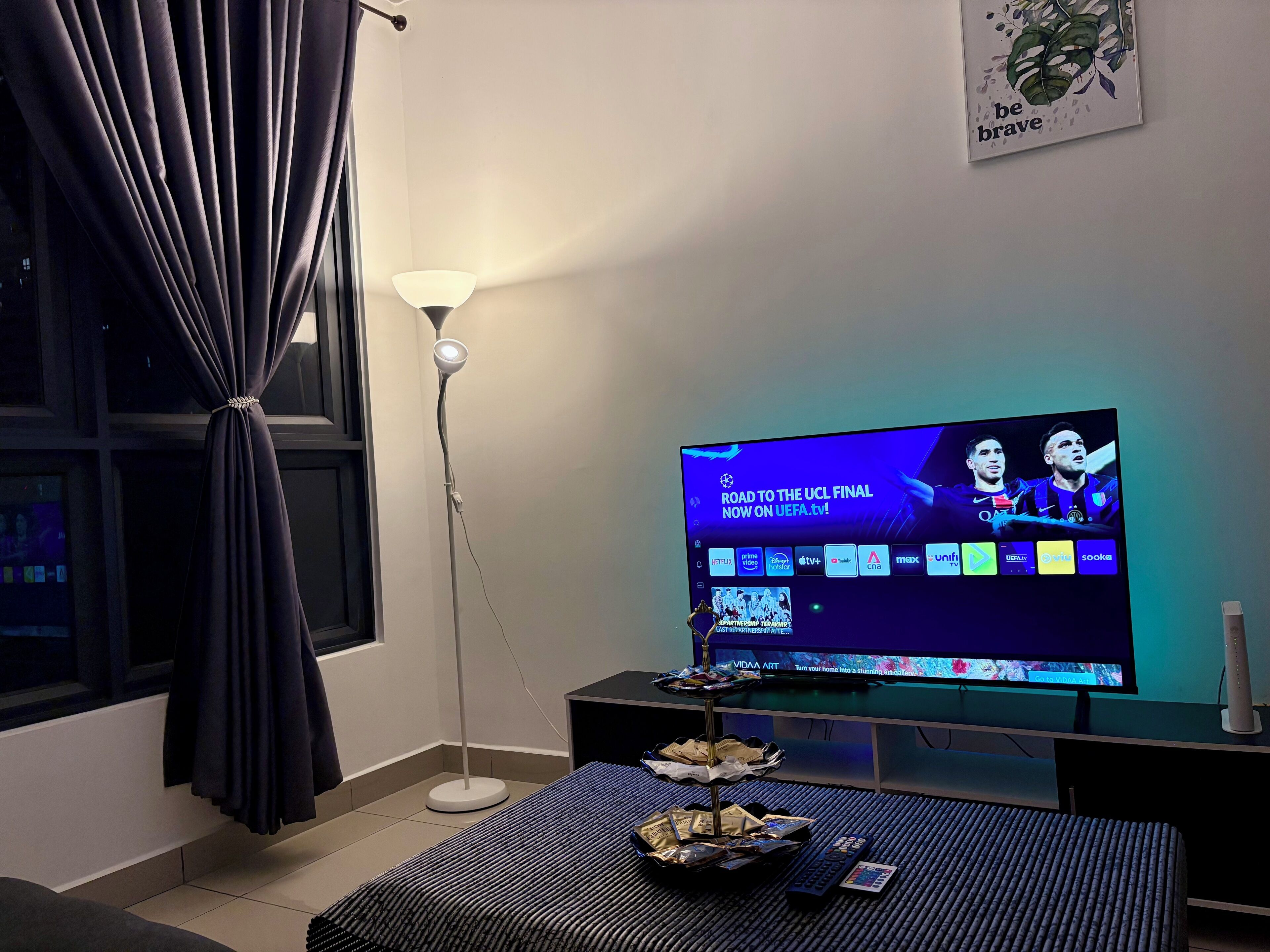 Una habitación con televisión mostrando un canal deportivo, una lámpara de pie, una mesita con aperitivos y una ventana con cortinas.