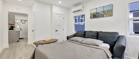 Un dormitorio con una cama, una mesita, un sofá y una kitchenette al fondo.
