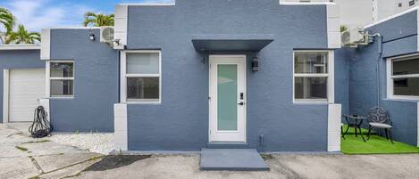 A modern building with a blue facade, a white door, and a small patio with two chairs.