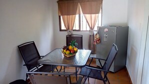 Un pequeño comedor con una mesa de vidrio, dos sillas, un refrigerador y una ventana con cortinas.