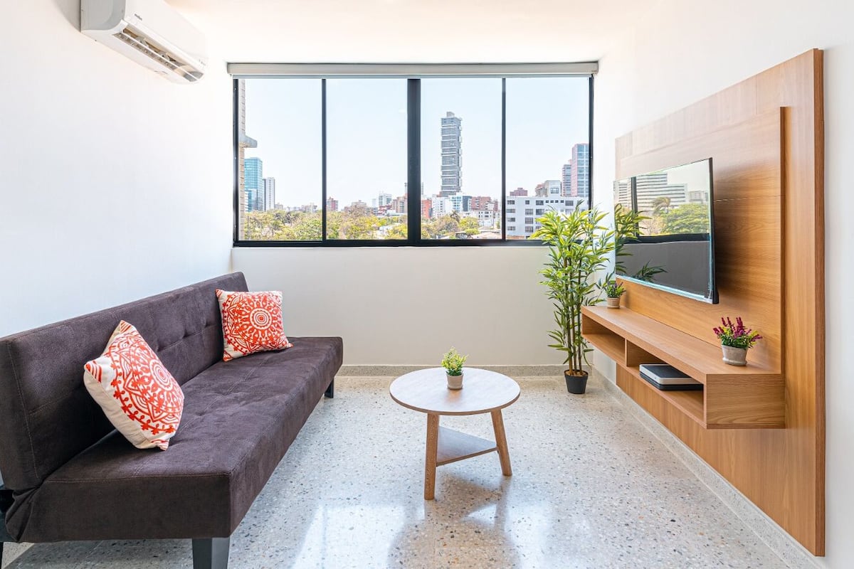 A modern living room with a dark brown sofa, two red patterned pillows, a round wooden coffee table, and a TV mounted on a wooden wall.