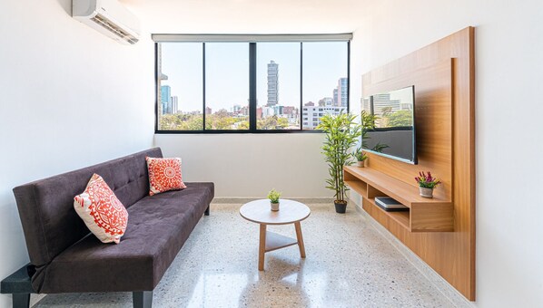 Una sala de estar moderna con un sofá marrón oscuro, dos almohadones rojos estampados, una mesa de centro redonda de madera y un televisor montado en una pared de madera.