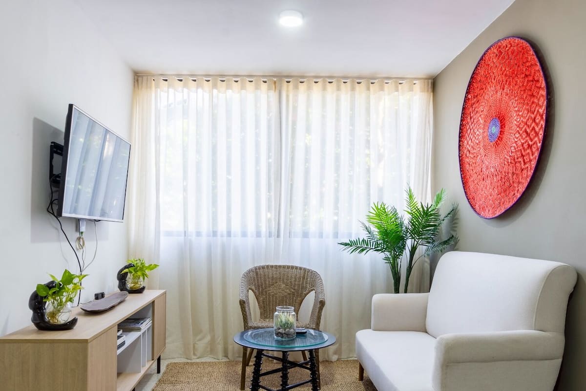 A living room with a white sofa, a glass coffee table, a wicker chair, a flat-screen TV mounted on the wall, and a large red circular artwork.