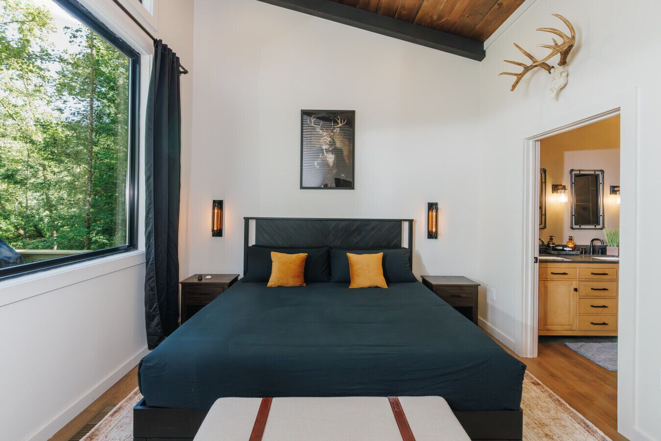 A bedroom with a large bed, two bedside tables, a nightstand, a window with curtains, a framed picture on the wall, and a wooden ceiling beam.