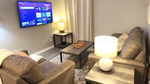 Uma sala de estar com televisão de tela plana na parede exibindo a interface de um serviço de streaming. Há um sofá, uma mesa de centro e um abajur sobre a mesa.