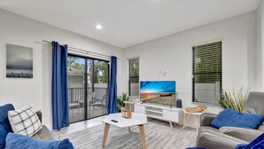 Ein modernes Wohnzimmer mit Flachbildfernseher, einer Couch, einem Couchtisch und einem Balkon mit blauen Vorhängen.