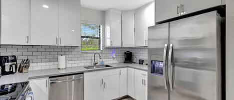 A modern kitchen with white cabinets, stainless steel appliances, and a window with blinds.