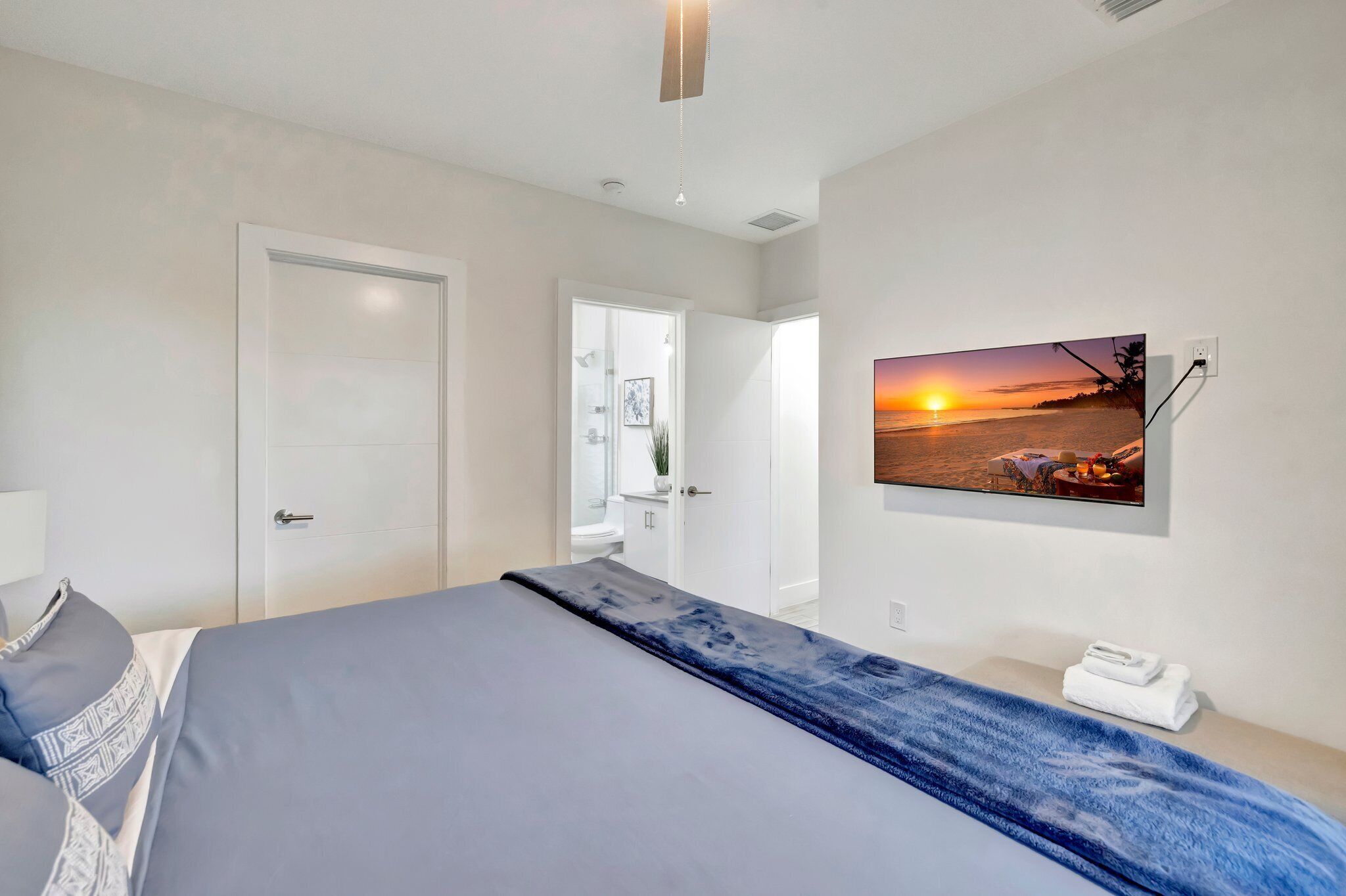 Une chambre moderne avec un lit, un téléviseur fixé au mur qui diffuse une image de coucher de soleil, et une porte donnant accès à une salle de bain.