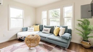 Ein Wohnzimmer mit grauem Sofa, einem gewebten Hocker und einem großen Fenster.