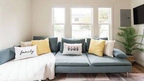 Ein modernes Wohnzimmer mit einem grauen Sofa, mehreren Kissen, einem 'family'-Kissen und einem großen Fenster mit Blick ins Freie.