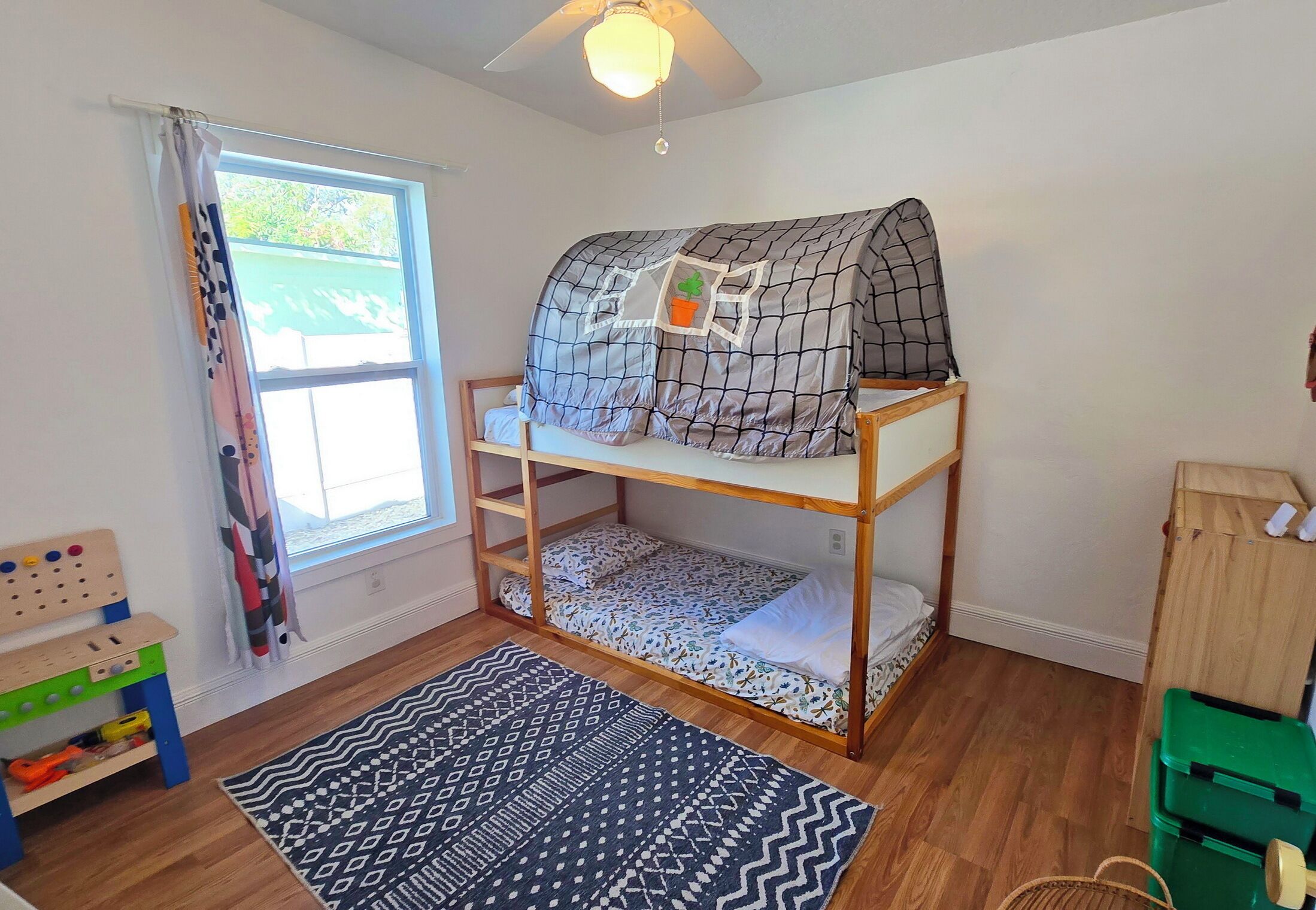 Un dormitorio infantil con literas, una ventana con cortinas, una mesa de juegos de madera y una alfombra estampada.