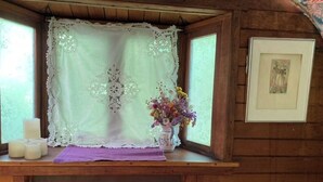 A wooden cabin with a window adorned with lace curtains, a vase of flowers, and candles on a wooden ledge.
