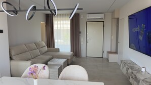 Una sala de estar moderna con un sofá seccional color beige, una mesa de centro de mármol y un jarrón decorativo con flores. Se observa un televisor de pantalla plana montado en la pared, una puerta con picaporte y una unidad de aire acondicionado encima.