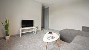 Ein modernes Wohnzimmer mit einem Flachbildfernseher auf einem weißen Schrank, einem kleinen Beistelltisch mit einer Pflanze, einem weißen Couchtisch mit Zeitschriften und einem grauen Sofa.
