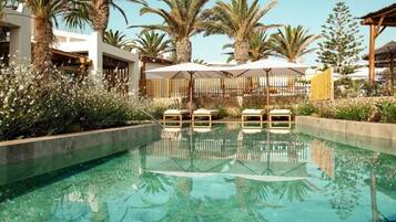 A clear swimming pool with lounge chairs and umbrellas, surrounded by palm trees and a wooden fence.