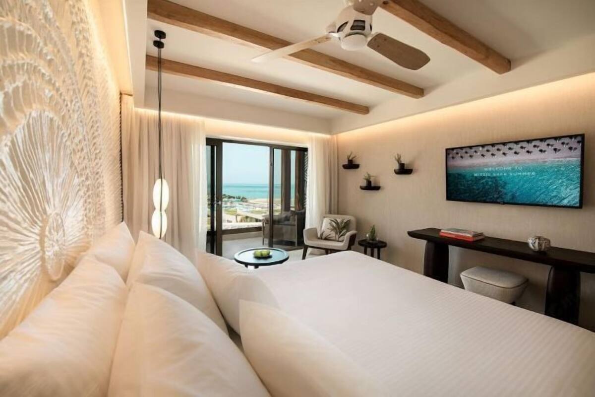 A hotel room with a bed, a desk with a TV, a chair, and a view of the sea.