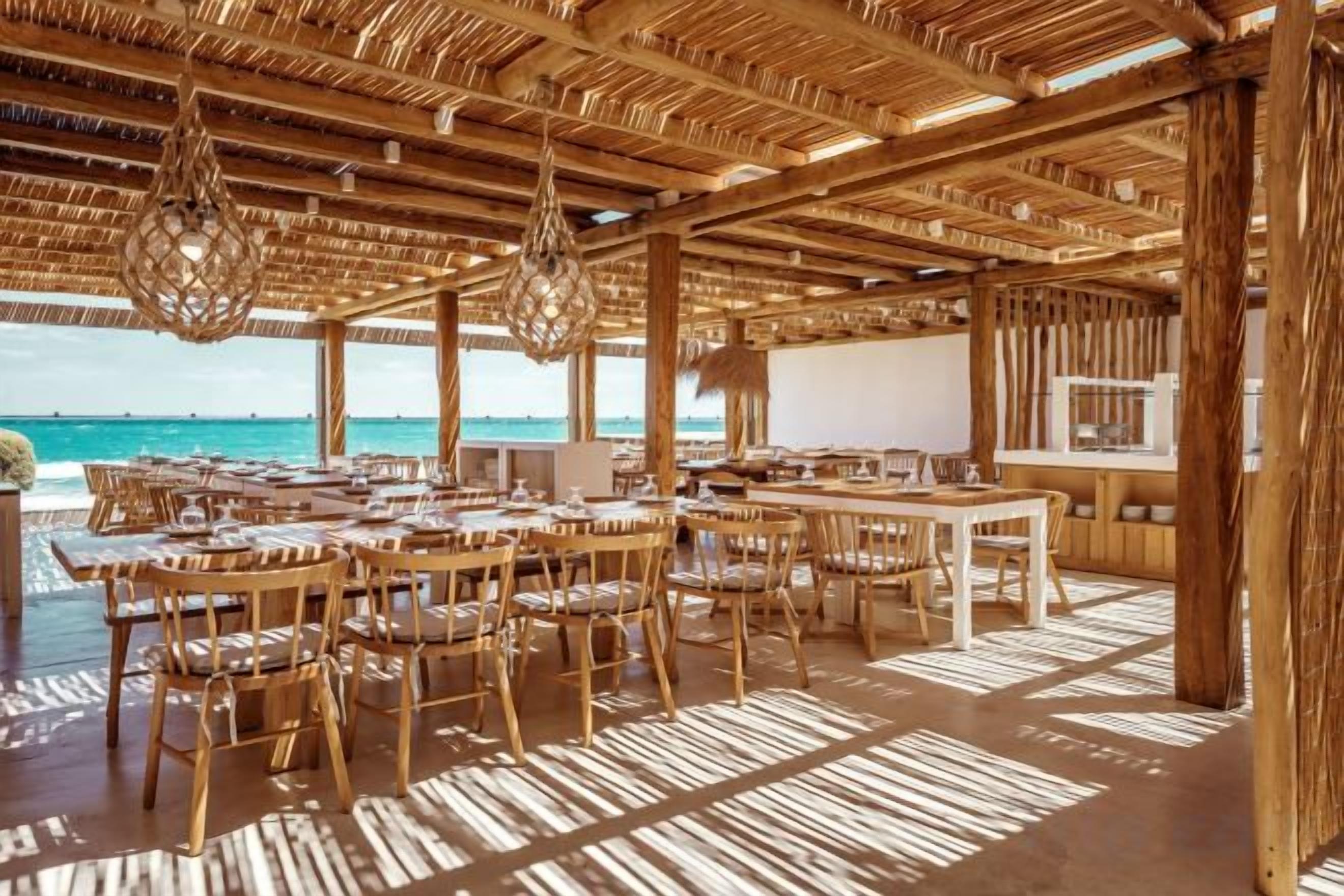 An outdoor dining area with wooden tables and chairs, a thatched roof, and a view of the ocean.