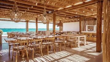 An outdoor dining area with wooden tables and chairs, a thatched roof, and a view of the ocean.