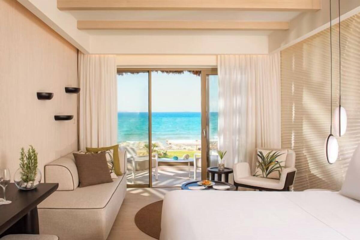 A modern hotel room with a large bed, a seating area with a view of the beach, and a balcony with a table and chairs.