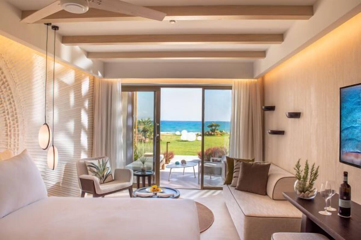A modern hotel room with a large bed, a seating area with a view of the beach, and a balcony with a glass door.