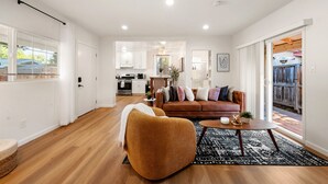 Ein modernes Wohnzimmer mit einem braunen Sofa, einem Couchtisch und einem schwarzen Teppich. Im Hintergrund befindet sich eine Küche mit weißen Schränken und einem Essbereich.