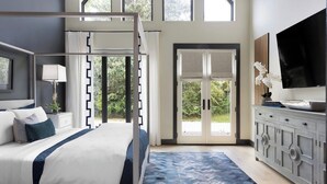 Un dormitorio con cama, mesita de noche, cómoda, televisor y un amplio ventanal con vista a zonas verdes.
