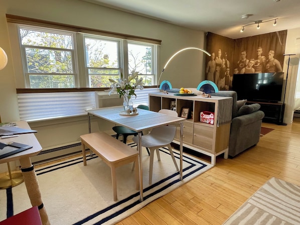 A modern living room with a dining area, a flat-screen TV, and a large window with blinds.
