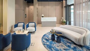 Eine moderne Hotelhalle mit einer Rezeption, einer Sitzecke mit Sofas und Sesseln sowie einem großen Fenster mit durchscheinenden Vorhängen.
