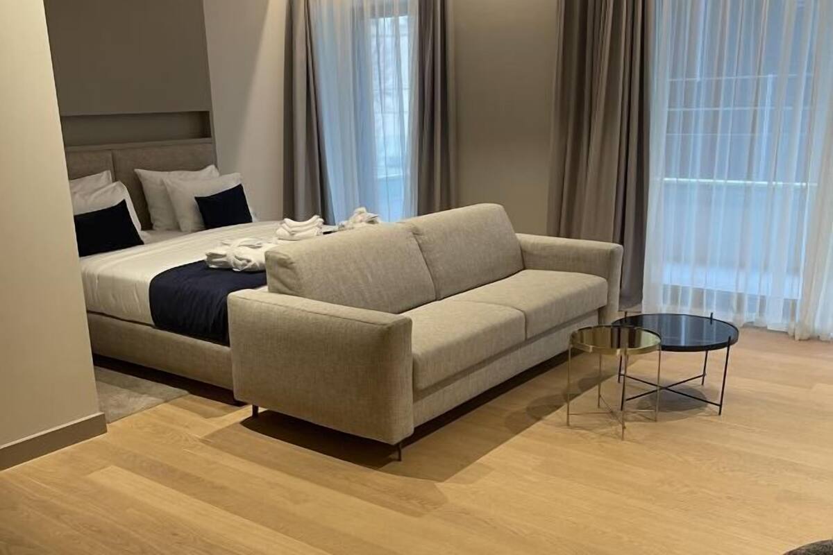 Una habitación de hotel moderna con una cama, un sofá color beige y una pequeña mesa redonda.