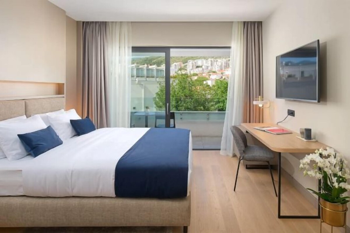 Habitación de hotel con cama, escritorio, silla y vistas a edificios a través de un ventanal.