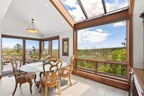 Enjoy meals in a dining area with a large round table and ornate chairs, surrounded by expansive windows offering a panoramic view of the lush landscape.
