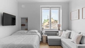 A modern bedroom with a bed, a grey sofa, a small table, and a window with a view of buildings.