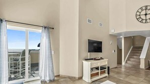 A spacious living area with a sliding glass door leading to a balcony, a flat-screen TV on a white console, and a staircase in the background. - Ocean View, 2 Primary Suites, Heated Pool, Beach (Cape Canaveral)