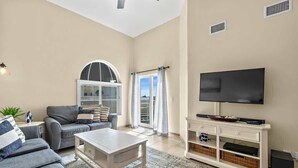 A living room with a grey sofa, a white coffee table, a flat-screen TV on a white stand, and a large window with curtains. - Ocean View, 2 Primary Suites, Heated Pool, Beach (Cape Canaveral)