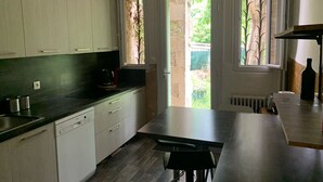 A kitchen with white cabinets, a black countertop, a dishwasher, and a table with chairs. There are windows with views of greenery outside. - Large villa 10 minutes from the Nîmes Arena (Nîmes)