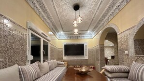 A traditional Moroccan interior with a long wooden dining table, striped sofas, intricate tile patterns, and a large flat-screen TV mounted on the wall. - Essaouira countryside, villa, swimming pool, 2 hectares, olive trees, mountain view, .... (essaouira)