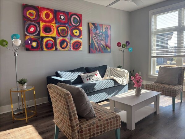 Uma sala de estar moderna com um sofá, duas poltronas, uma mesa de centro e um abajur. Há duas pinturas na parede e uma janela com persianas.