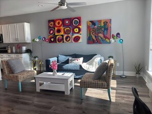 Sala de estar com sofá, duas poltronas, mesa de centro e ventilador de teto. Há duas pinturas na parede e um vaso com planta no chão.