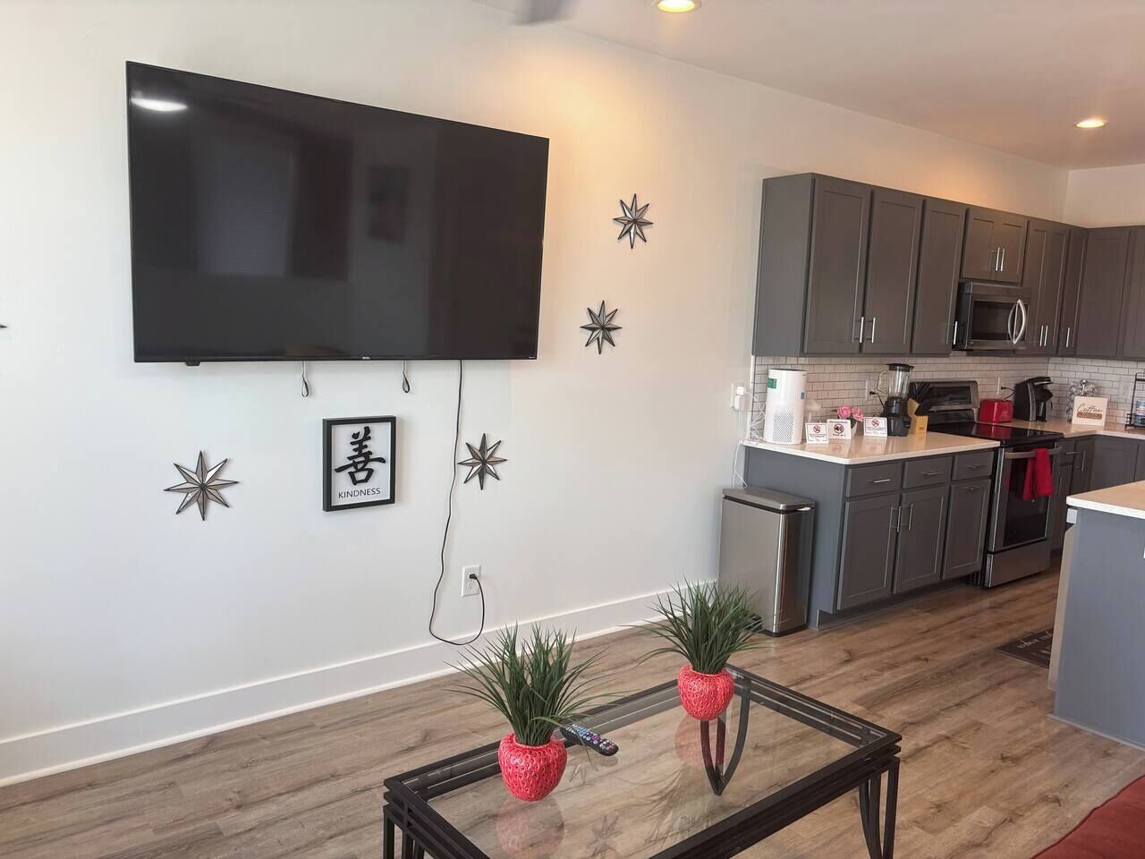 A modern kitchen with grey cabinets, a microwave, and a stove. There is a flat-screen TV mounted on the wall and a small trash bin.